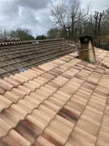 Rénovation de toiture: tuiles terre cuite posées, structure en lattes bois exposée, cheminée métallique et antenne TV sous ciel gris.