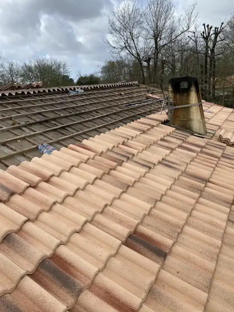 Rénovation de toiture: tuiles terre cuite posées, structure en lattes bois exposée, cheminée métallique et antenne TV sous ciel gris.