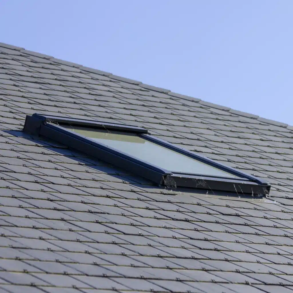 Fenêtre de Toit sur Toiture en Ardoise | Pose Velux Fenêtre de toit Velux installée sur une toiture en ardoise avec crochets sous un ciel bleu clair.